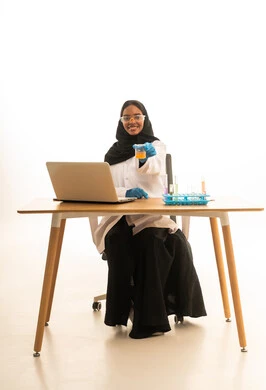 بورتريه لطالبة عربيه خليجيه سعوديه ترتدي العباءه والحجاب والمعطف الابيض ونظارات الحماية تستخدم جهاز الحاسوب المحمول تجلس امام طاولة الاختبارات وأنابيب العينات والتحاليل الكيميائية، أساليب التعلم الفعال بالاستفادة من التقنيات والأجهزة التكنولوجية، استخدام الأدوات والوسائل التعليمية التجريبية،خلفية بيضاء