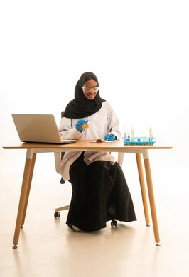 أساليب التعلم الفعال بالاستفادة من التقنيات والأجهزة التكنولوجية، استخدام الأدوات والوسائل التعليمية التجريبية، بورتريه لطالبة عربيه خليجيه سعوديه ترتدي العباءه والحجاب والمعطف الابيض ونظارات الحماية تستخدم جهاز الحاسوب المحمول تجلس امام طاولة الاختبارات وأنابيب العينات والتحاليل الكيميائية، خلفية بيضاء