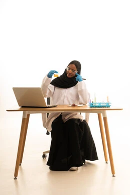 استخدام الأدوات والوسائل التعليمية التجريبية، أساليب التعلم الفعال بالاستفادة من التقنيات والأجهزة التكنولوجية، بورتريه لطالبة عربيه خليجيه سعوديه ترتدي العباءه والحجاب والمعطف الابيض ونظارات الحماية تستخدم جهاز الحاسوب المحمول تجلس امام طاولة الاختبارات وأنابيب العينات والتحاليل الكيميائية، خلفية بيضاء