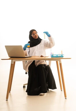 بورتريه لطالبة عربيه خليجيه سعوديه ترتدي العباءه والحجاب والمعطف الابيض ونظارات الحماية تستخدم جهاز الحاسوب المحمول تجلس امام طاولة الاختبارات وأنابيب العينات والتحاليل الكيميائية، أساليب التعلم الفعال بالاستفادة من التقنيات والأجهزة التكنولوجية، استخدام الأدوات والوسائل التعليمية التجريبية،خلفية بيضاء