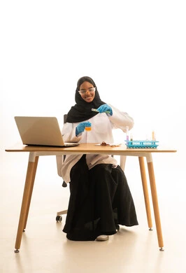التطور والبحث وإجراء التفاعلات الكيميائية, استخدام الأدوات والوسائل التعليمية التجريبية، بورتريه لطالبة عربيه خليجيه سعوديه ترتدي العباءه والحجاب والمعطف الابيض ونظارات الحماية تستخدم جهاز الحاسوب المحمول تجلس امام طاولة الاختبارات وأنابيب العينات والتحاليل الكيميائية، خلفية بيضاء