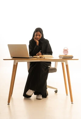 The use of modern technologies, the concept of homeschooling, a portrait of a Gulf Arab Saudi woman wearing an abaya and hijab sitting at a study desk holding a book, using a computer to complete her assignments, the role of technology in changing traditional teaching methods, white background.