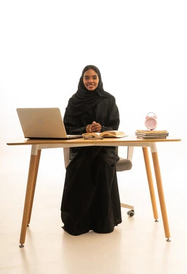 دور التكنولوجيا في تغيير أساليب التعليم التقليدية,مفهوم التعليم المنزلي, بورتريه لامراه عربية خليجية سعودية ترتدي العباءة والحجاب تجلس على المكتب الدراسي تمسك بيدها كتاب , استخدام جهاز الحاسوب في حل واجباتها , خلفية بيضاء.