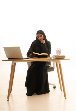 The concept of homeschooling, a portrait of a Gulf Arab Saudi woman wearing an abaya and hijab sitting at a study desk holding a book, using a computer to complete her homework, the role of technology in changing traditional teaching methods, white background.
