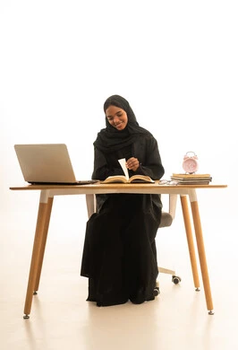 The arrival of exam and study time, the use of technology and modern techniques, the development of curricula and educational materials in Saudi Arabia, making the most of time for studying and reviewing, a portrait of a Gulf Arab Saudi university student wearing an abaya and hijab in front of an alarm clock, books, and a laptop, with a white background.