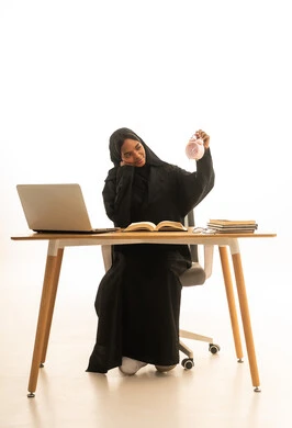 The arrival of exam and study time, the use of technology and modern techniques, the development of curricula and educational materials in Saudi Arabia, making the most of time for studying and reviewing, a portrait of a Gulf Arab Saudi university student wearing an abaya and hijab in front of an alarm clock, books, and a laptop, with a white background.