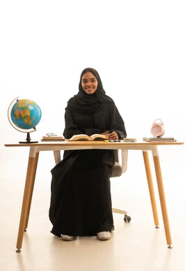 ايماءات التفوق والاجتهاد , مراجعة الدروس اليومية ،  مفهوم البحث والاستكشاف حول العالم, بورتريه لطالبة عربية خليجية سعودية ترتدي العباءة والحجاب تجلس على الطاولة امام  مجسم الكرة الارضية والكتب ، خلفية بيضاء
