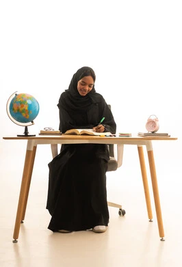 The concept of research and exploration around the world, reviewing daily lessons with gestures of excellence and diligence, a portrait of an Arab Gulf Saudi student wearing an abaya and hijab sitting at a table in front of a laptop and a globe model and books, with a white background.