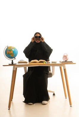 استخدام التكنولوجيا والتقنيات الحديثة، تحديد هدف معين ، قدوم وقت الامتحانات والمذاكرات، مفهوم البحث والاستكشاف حول العالم, بورتريه لطالبة عربية خليجية سعودية ترتدي العباءة والحجاب ونظارات الفى ار تجلس على الطاولة امام مجسم الكرة الارضية والكتب ، خلفية بيضاء