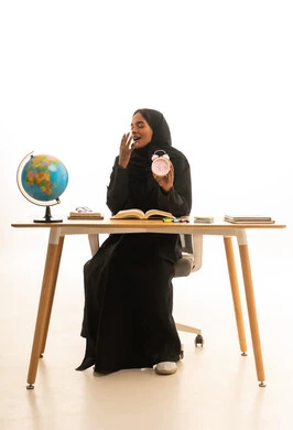 Expressions of drowsiness and the need for sleep, the arrival of exam and study time, the concept of excellence and diligence, the concept of research and exploration around the world, a portrait of an Arab Gulf Saudi student wearing an abaya and hijab sitting at a table in front of a globe model and books, with a white background.