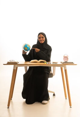 الاشارة الى الخريطة, قدوم وقت الامتحانات والمذاكرات، مفهوم التفوق والاجتهاد , مفهوم البحث والاستكشاف حول العالم, بورتريه لطالبة عربية خليجية سعودية ترتدي العباءة والحجاب تجلس على الطاولة امام مجسم الكرة الارضية والكتب ، خلفية بيضاء