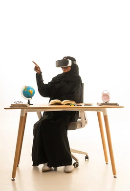 Wearing virtual reality glasses, setting a specific goal, the arrival of exam time and studying, the concept of research and exploration around the world, a portrait of an Arab Gulf Saudi student wearing an abaya and hijab and VR glasses sitting at a table in front of a globe model and books, white background.