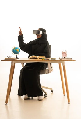 ارتداء نظارات الواقع الافتراضي، تحديد هدف معين ، قدوم وقت الامتحانات والمذاكرات، مفهوم البحث والاستكشاف حول العالم, بورتريه لطالبة عربية خليجية سعودية ترتدي العباءة والحجاب ونظارات الفى ار تجلس على الطاولة امام مجسم الكرة الارضية والكتب ، خلفية بيضاء