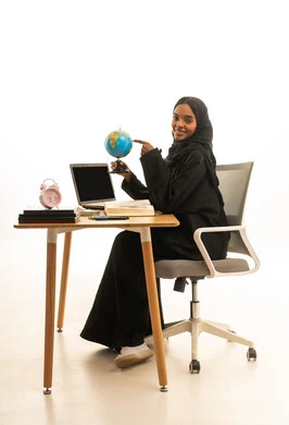 مفهوم البحث والاستكشاف حول العالم ، استخدام التكنولوجيا والتقنيات الحديثة، قدوم وقت الامتحانات والمذاكرات، بورتريه لطالبة عربية خليجية سعودية ترتدي العباءة والحجاب تجلس على الطاولة امام مجسم الكرة الارضية والكتب والحاسوب المحمول ، خلفية بيضاء