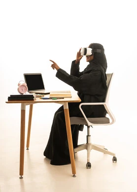 The arrival of exam and study time, the use of technology and modern techniques, setting a specific goal, the concept of research and exploration around the world, a portrait of a Gulf Arab Saudi student wearing an abaya and hijab and VR glasses sitting at a table in front of a globe model, books, and a laptop, with a white background.