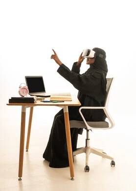 The use of technology and modern techniques, setting a specific goal, the arrival of exam time and studying, the concept of research and exploration around the world, a portrait of an Arab Gulf Saudi student wearing an abaya and hijab and VR glasses sitting at a table in front of a globe model, books, and a laptop, with a white background.