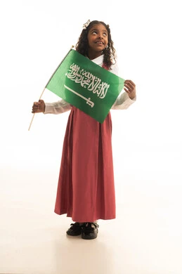 الفخر والاعتزاز بالوطن، وإحياء ذكرى اليوم الوطني السعودي في المدارس، بورتريه لطالبه عربية خليجية سعودية ترتدي الزي المدرسي والحقيبة المدرسية، تلميذه مجتهدة ترتدي الزي المدرسي الوردي تحمل بيدها علم المملكة العربية السعودية، خلفية بيضاء.