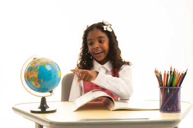 The concept of research and exploration around the world, the use of tools and aids, and the quality of effective learning methods, a close portrait of a Saudi Gulf Arab student wearing a school uniform sitting at a desk pointing to a globe model, studying school lessons, with a white background.