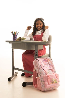 Raising hands with expressions of enthusiasm and joy, studying school lessons, using tools and aids, and the quality of effective learning methods, a close portrait of a Saudi Gulf Arab student wearing a school uniform sitting at a desk with a globe model beside her, white background.