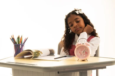 الارهاق والملل خلال الدوام، الشعور بتأخر الوقت والحاجة للنوم، موسم العودة إلى المدارس، بورتريه لطالبة عربية خليجية سعودية ترتدي الزي المدرسي  تجلس في مقعدها تقوم بالنظر إلى ساعة المنبه، قرطاسية مدرسية على الطاولة المكتبية، خلفية بيضاء