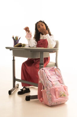 The feeling of time running late and the need for sleep, fatigue and exhaustion during work hours, the back-to-school season, a portrait of a Saudi Gulf Arab student wearing a school uniform sitting in her seat looking at the alarm clock, school stationery on the desk, white background.