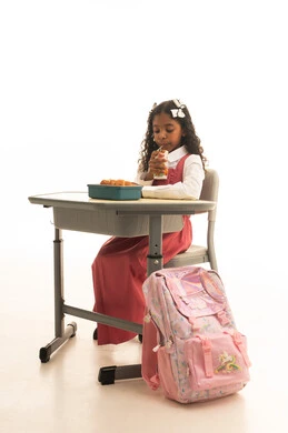 الاستمتاع بتناول الأطعمة الصحية في وقت الفسحة المدرسية، موسم العودة للمدارس، بورتريه مقرب لطالبة عربيه خليجيه سعودية ترتدي الزي المدرسة تجلس على الطاوله المكتبية، تناول وجبة الفطور الصحية في الفسحة المدرسية، خلفية بيضاء