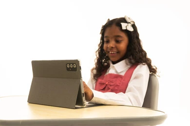 The concept of distance learning, attending online classes via the internet, using modern technology, a portrait of an Arab Gulf Saudi student wearing a school uniform sitting at a study table working on a tablet with expressions of joy, white background.