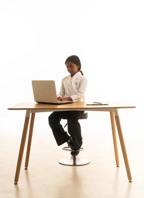 تدوين نتائج الفحوصات، الهدف والطموح المستقبلي، استخدام الاجهزة التقنية الحديثة، بورتريه مقرب لبنت  عربية خليجية سعودية  ترتدي المعطف الأبيض تجلس خلف المكتب تعمل على الحاسوب المحمول، فتاة سعودية  تقوم بإعداد التقارير، صورة لكامل الجسم، خلفية بيضاء