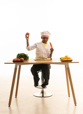 Delicious recipes and the joy of preparing them, a portrait of a Saudi Gulf Arab girl wearing a chef's outfit with a white hat, holding various colored vegetables in her hands and comparing them with gestures of discovery and happiness, white background.