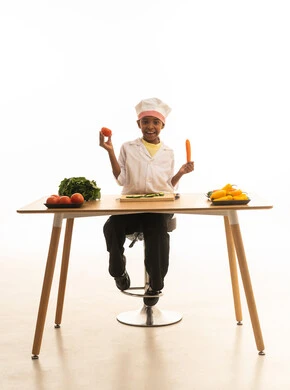 الوصفات الشهية و المتعة في تحضيرها , بورتريه لطفلة عربية خليجيه سعودية ترتدي زي الشيف مع قبعته البيضاء, تمسك بيديها الخضروات المختلفة الألوان وتقارن بينها بإيماءات الاكتشاف والسعادة, خلفية بيضاء