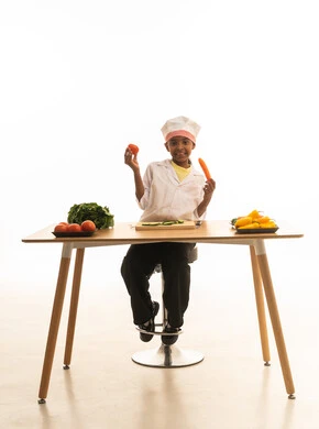 المتعة في تحضير الوصفات الشهية, بورتريه لطفلة عربية خليجيه سعودية ترتدي زي الشيف مع قبعته البيضاء, تمسك بيديها الخضروات المختلفة الألوان وتقارن بينها بإيماءات الاكتشاف والسعادة, خلفية بيضاء