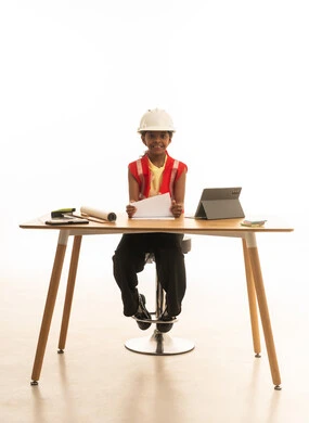 تجسيد تجربة العمل كمهندسة معمارية، تجريب الأطفال للمهن المستقبلية منذ الصغر، بورتريه لبنت عربية خليجية سعودية ترتدي سترة الحماية تجلس خلف المكتب وتتابع مخطط الإعمار بإيماءات التركيز، خلفية بيضاء