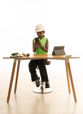 Children experimenting with future professions from a young age, embodying the experience of working as an architect, a portrait of a Saudi Gulf Arab girl wearing a safety vest sitting behind a desk and following the construction plan with focused gestures, white background.