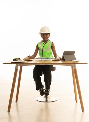 The use of modern technology in professions, allowing children to experiment with future careers from a young age, embodying the experience of working as an architect, a portrait of a Saudi Gulf Arab girl wearing a safety vest sitting behind a desk and following the construction plan with focused gestures, white background.