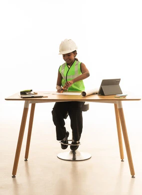 تجسيد تجربة العمل كمهندسة معمارية، تجريب الأطفال للمهن المستقبلية منذ الصغر، بورتريه لبنت عربية خليجية سعودية ترتدي سترة الحماية تجلس خلف المكتب وتتابع مخطط الإعمار بإيماءات التركيز، خلفية بيضاء