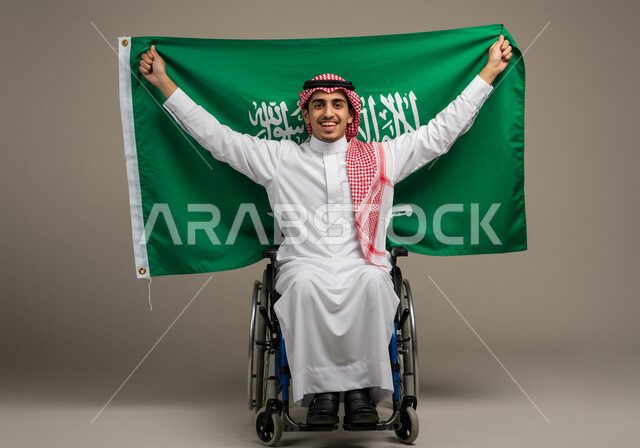 مشاركة ذوي الاحتياجات الخاصة بالاحتفال بالمناسبات والأعياد الوطنية، إحياء ذكرى اليوم الوطني السعودي 23 سبتمبر، تقديم الولاء والحب للوطن، بورتريه لشاب عربي خليجي سعودي يرتدي الثوب التقليدي والشماغ يجلس على الكرسي المتحرك يحمل علم المملكة، خلفية رمادية