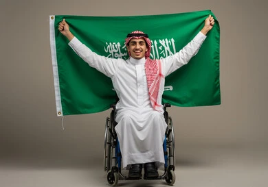 Participating with people with special needs in celebrating national occasions and holidays, commemorating Saudi National Day, September 23. Expressing loyalty and love for the homeland. Portrait of a young Saudi Arabian Gulf man wearing a traditional thobe and shemagh, sitting in a wheelchair holding the Kingdom's flag, gray background.