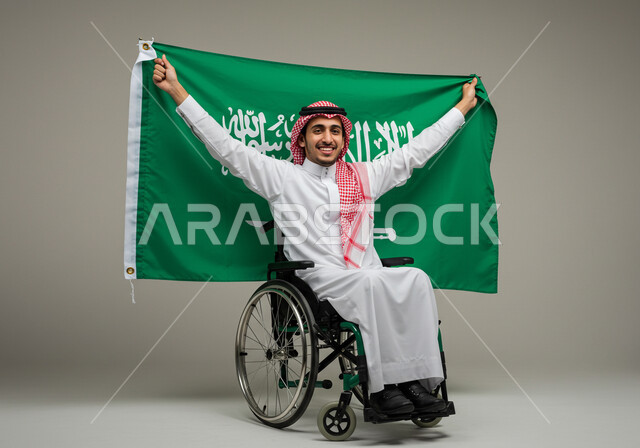 مشاركة ذوي الاحتياجات الخاصة بالاحتفال بالمناسبات والأعياد الوطنية، إحياء ذكرى اليوم الوطني السعودي 23 سبتمبر، تقديم الولاء والحب للوطن، بورتريه لشاب عربي خليجي سعودي يرتدي الثوب التقليدي والشماغ يجلس على الكرسي المتحرك يحمل علم المملكة، خلفية رمادية