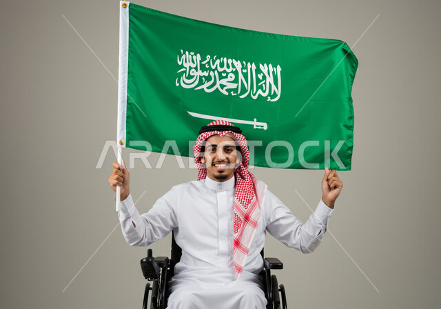 مشاركة ذوي الاحتياجات الخاصة بالاحتفال بالمناسبات والأعياد الوطنية، إحياء ذكرى اليوم الوطني السعودي 23 سبتمبر، تقديم الولاء والحب للوطن، بورتريه لشاب عربي خليجي سعودي يرتدي الثوب التقليدي والشماغ يجلس على الكرسي المتحرك يحمل علم المملكة، خلفية رمادية