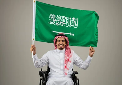 Participating with people with special needs in celebrating national occasions and holidays, commemorating Saudi National Day, September 23. Expressing loyalty and love for the homeland. Portrait of a young Saudi Arabian Gulf man wearing a traditional thobe and shemagh, sitting in a wheelchair holding the Kingdom's flag, gray background.