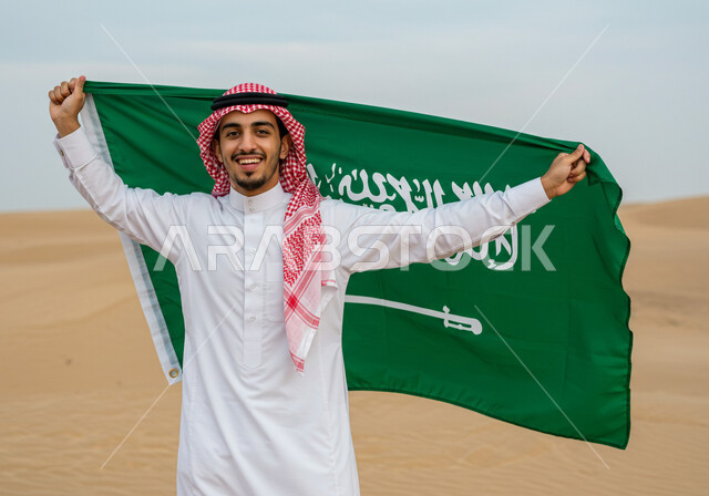 الاحتفال بالمناسبات والأعياد الوطنية في الطبيعة، يوم العلم 11 مارس، اليوم الوطني السعودي 23 سبتمبر، رجل عربي خليجي سعودي يرتدي الشماغ والثوب التقليدي يقف وسط الصحراء يحمل علم المملكة العريية السعودية لاحياء الذكرى 
