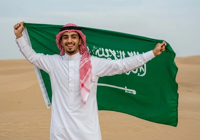 الاحتفال بالمناسبات والأعياد الوطنية في الطبيعة، يوم العلم 11 مارس، اليوم الوطني السعودي 23 سبتمبر، رجل عربي خليجي سعودي يرتدي الشماغ والثوب التقليدي يقف وسط الصحراء يحمل علم المملكة العريية السعودية لاحياء الذكرى 