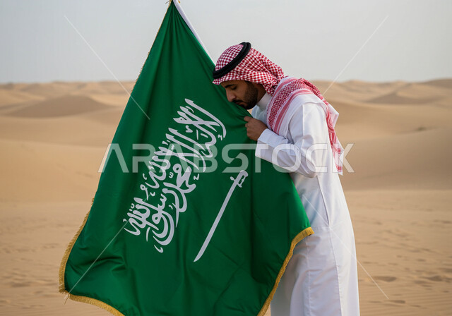 الاحتفال بالمناسبات والأعياد الوطنية في الطبيعة، يوم العلم 11 مارس، اليوم الوطني السعودي 23 سبتمبر، رجل عربي خليجي سعودي يرتدي الشماغ والثوب التقليدي يقف وسط الصحراء يقبل علم المملكة العريية السعودية لاحياء الذكرى 
