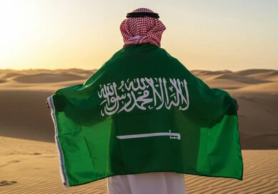 National events and holidays in nature, celebrating Flag Day on March 11, Saudi National Day on September 23, a photo from the back of a Saudi Arabian Gulf man wearing a traditional shemagh and thobe standing in the middle of the desert with the flag of the Kingdom of Saudi Arabia on his shoulders to commemorate the anniversary