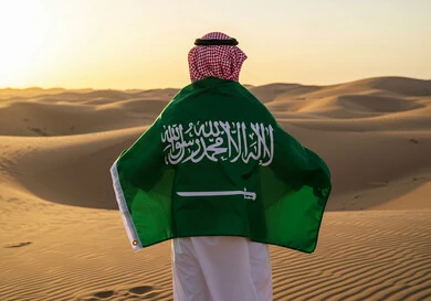 المناسبات والأعياد الوطنية في الطبيعة، الاحتفال بيوم العلم 11 مارس، اليوم الوطني السعودي 23 سبتمبر، صورة من الخلف لرجل عربي خليجي سعودي يرتدي الشماغ والثوب التقليدي يقف وسط الصحراء يضع علم المملكة العريية السعودية على كتفيه لاحياء الذكرى 