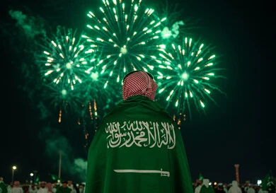 مشاهدة المفرقعات والألعاب النارية في المساء، صورة من الخلف لرجل عربي خليجي سعودي يرتدي الشماغ يضع علم المملكة العربية السعودية على كتفيه، الاحتفال بالمناسبات والأعياد الوطنية، يوم العلم 11 مارس، اليوم الوطني السعودي 23 سبتمبر، احياء الذكرى 95  للسعودية