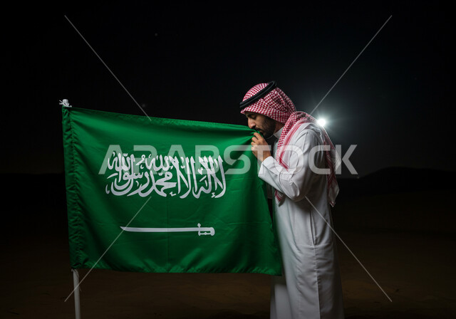 اليوم الوطني السعودي 23 سبتمبر، راية التوحيد وشعار الوطن، صورة مقربة من الجانب لرجل عربي خليجي سعودي يرتدي الشماغ والثوب التقليدي يقبل العلم بايماءات الفخر والاعتزاز، الاحتفال بالمناسبات والأعياد الوطنية، يوم العلم 11 مارس
