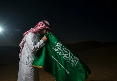 Saudi National Day, September 23, the banner of unification and the emblem of the nation, a close-up side view of a Saudi Arabian Gulf man wearing a traditional shemagh and thobe kissing the flag with gestures of pride and honor, celebrating national occasions and holidays, Flag Day, March 11