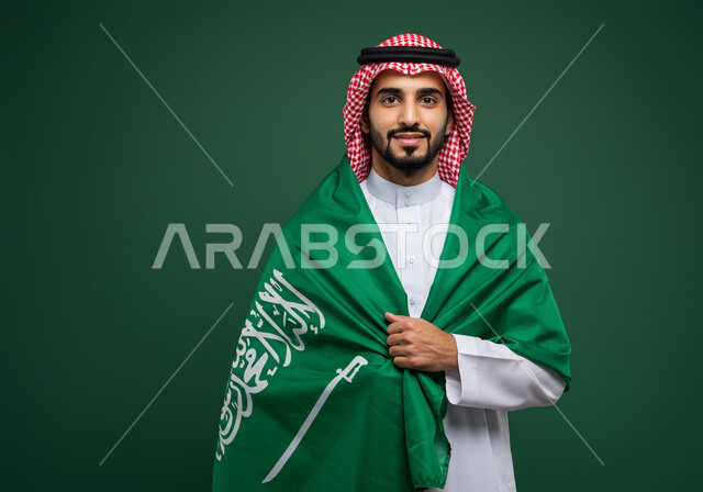 النظر الى الكاميرا بتعابير السرور والفخر بالانتماء للوطن، بورتريه رجل عربي خليجي سعودي مبتسم يرتدي الشماغ والثوب التقليدي يضع الراية على كتفه يحتفل باليوم الوطني السعودي 23 سبتمبر، يوم العلم 11 مارس، احتفالات ومناسبات وطنية في المملكة العربية السعودية، خلفية خضراء