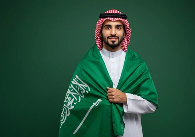 Looking at the camera with expressions of joy and pride in belonging to the homeland, portrait of a smiling Saudi Arabian Gulf man wearing a traditional shemagh and thobe, placing the flag on his shoulder, celebrating Saudi National Day, September 23, Flag Day, March 11, national celebrations and occasions in the Kingdom of Saudi Arabia, green background